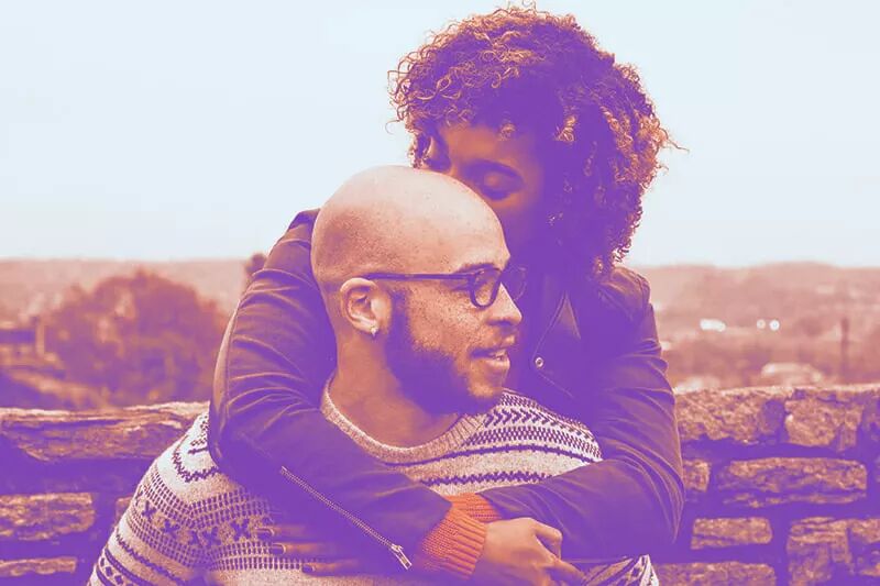 Woman standing behind a man who is sitting kissing his head while she has her arms wrapped around hi…